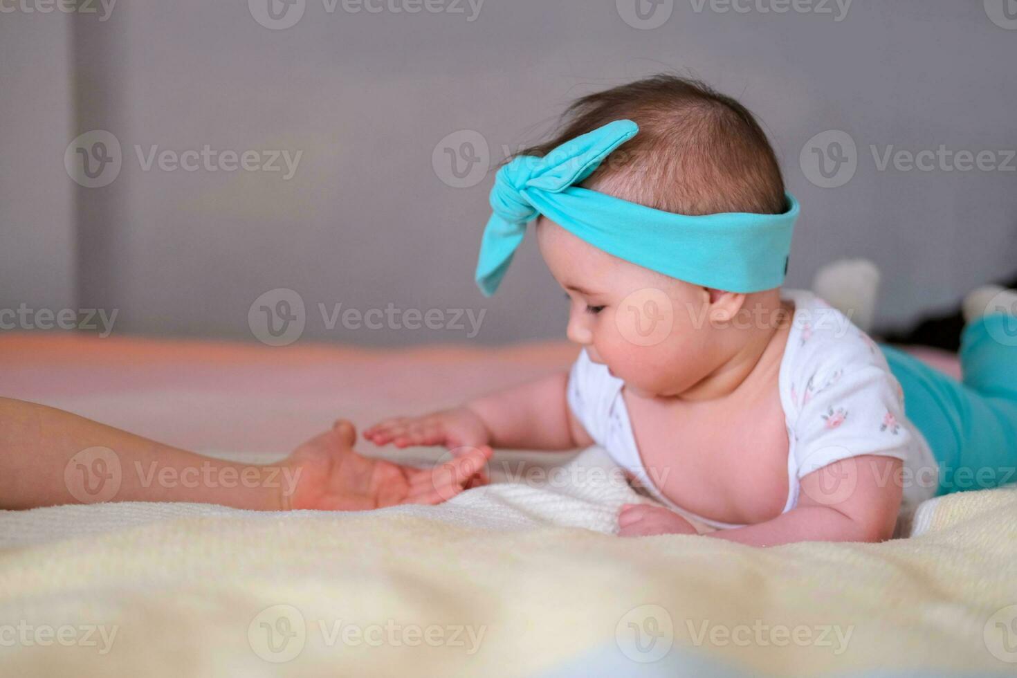 une 6 mois vieux bébé mensonges sur le sien estomac sur le lit et touche le sien sœurs main. une magnifique Bandeau pour une bébé. photo