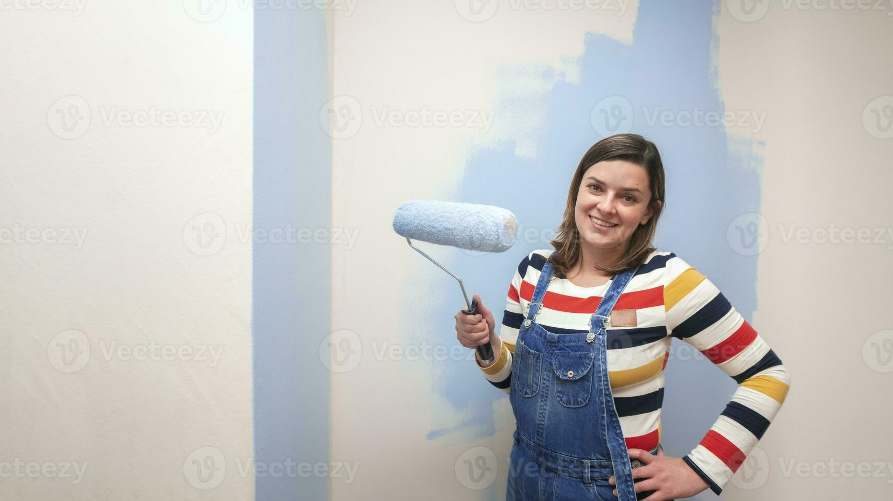 magnifique femme habillé dans salopette, souriant à caméra avec bleu peindre rouleau dans sa main contre à moitié peint blanc mur Contexte photo