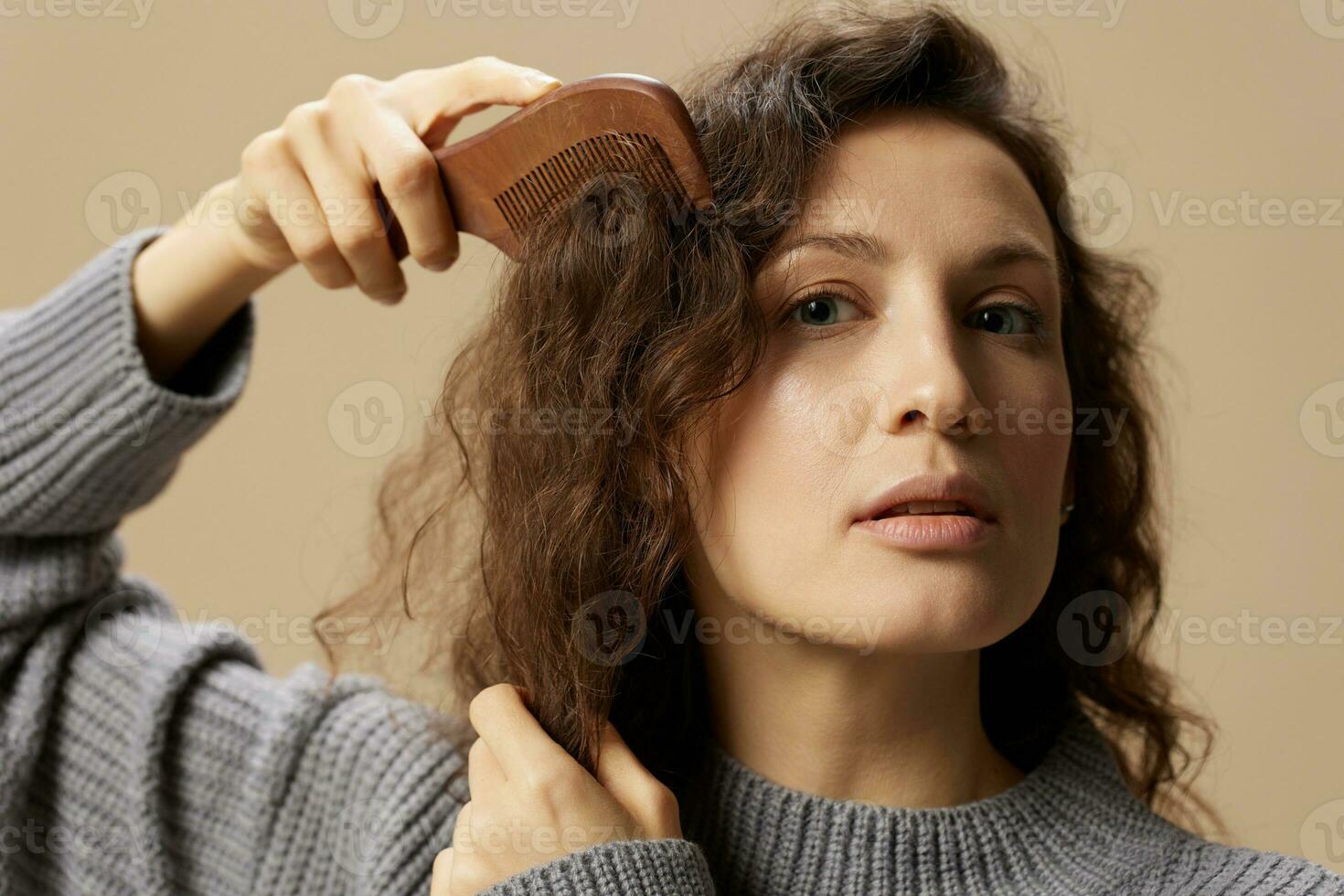 adorable jolie frisé magnifique femelle dans gris décontractée chandail avec brosse à cheveux peigne en bonne santé boucles posant isolé sur plus de beige pastel Contexte. problématique indiscipliné endommagé cheveux concept. copie espace photo