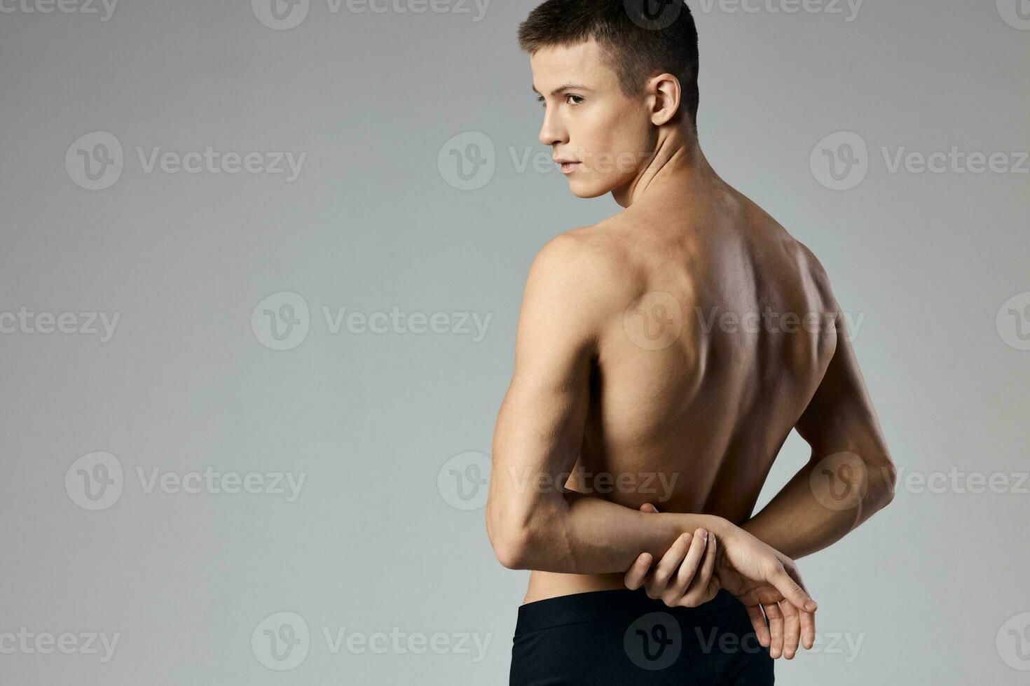 une homme avec pompé en haut muscles détient le sien mains derrière le sien retour sur une gris Contexte et derrière modèle photo