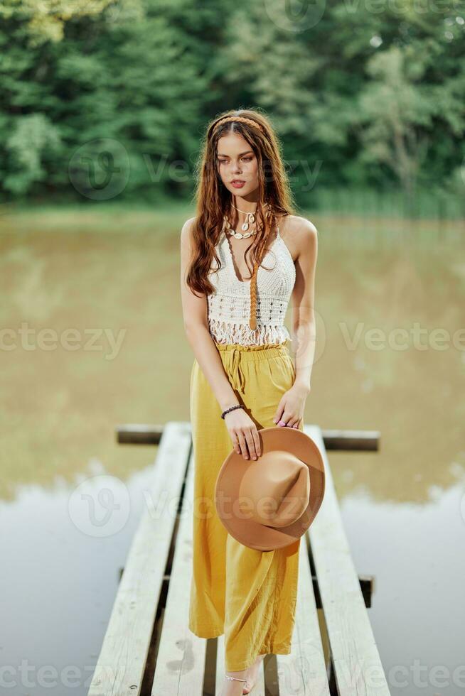 une Jeune femme dans une hippie Regardez et éco-robe dansant en plein air par le Lac portant une chapeau et Jaune un pantalon dans le été le coucher du soleil photo