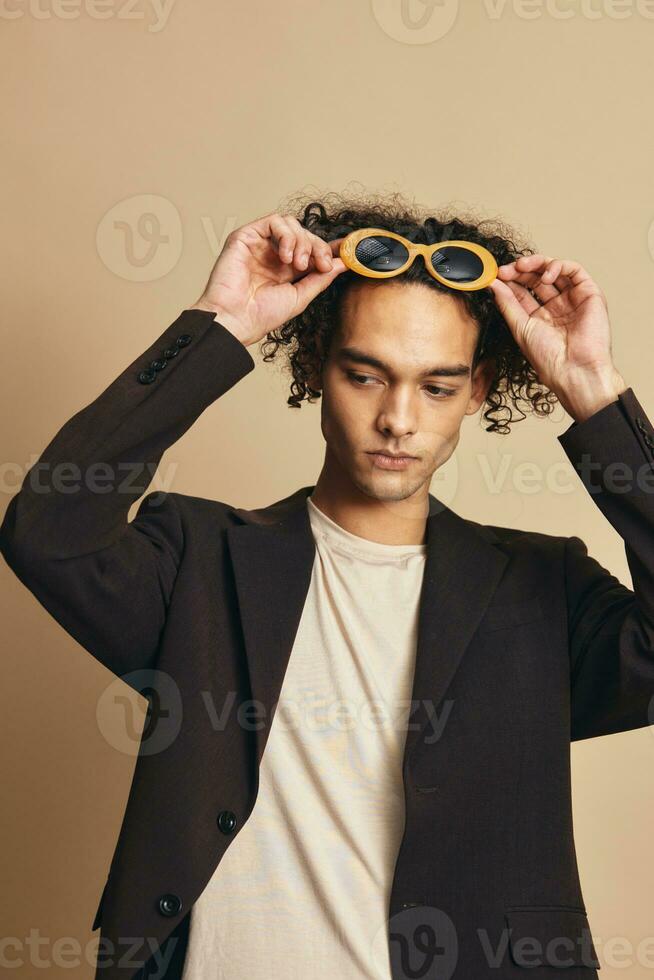 à la mode impressionnant bronzé frisé homme dans classique marron veste branché des lunettes de soleil posant isolé sur plus de beige pastel Contexte. mode Nouveau collection offre. rétro style concept. gratuit endroit pour un d photo