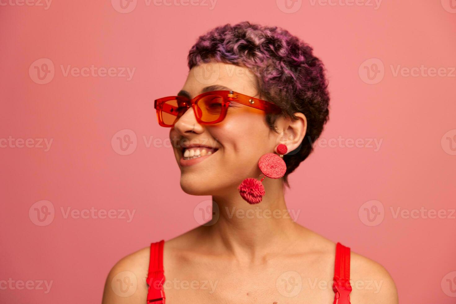 mode portrait de une femme avec une court la Coupe de cheveux dans coloré des lunettes de soleil avec inhabituel accessoires avec des boucles d'oreilles souriant sur une rose brillant Contexte photo