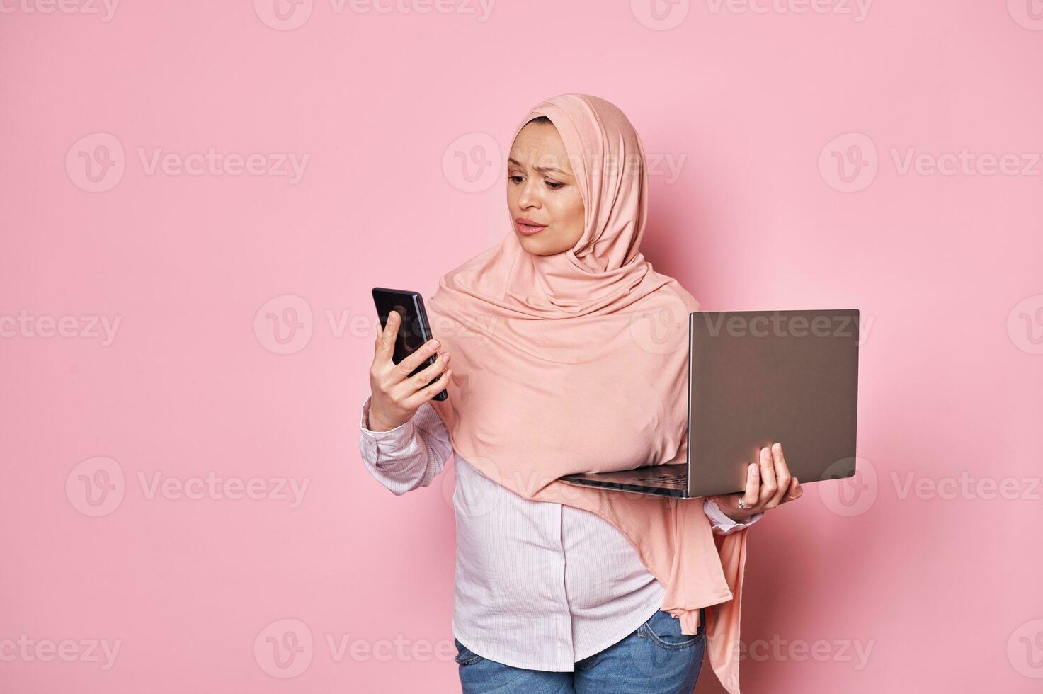 perplexe arabe musulman Enceinte femme dans hijab, à la recherche à sa téléphone intelligent, posant avec portable plus de rose Couleur Contexte photo