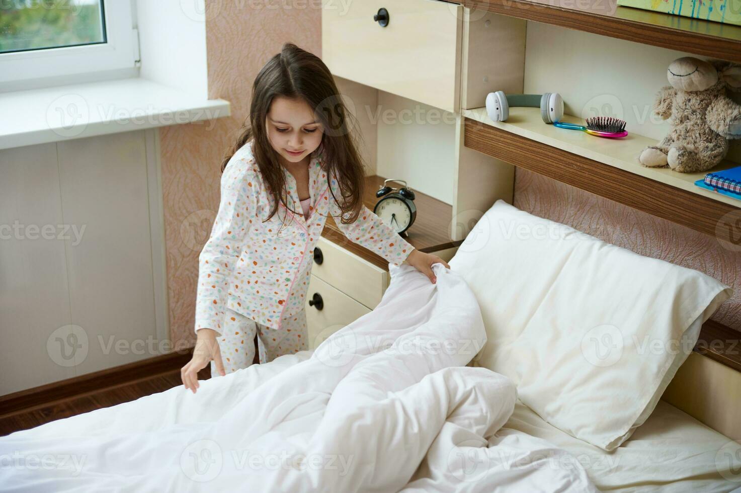 peu fille dans pyjamas se redresse lit feuilles et couette sur sa lit après en train de dormir et nettoie sa pièce dans le Matin photo