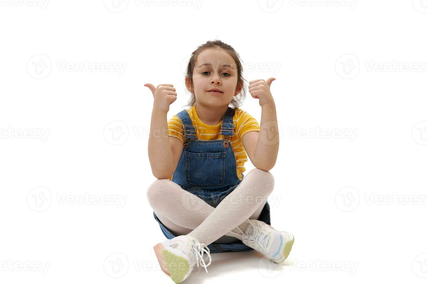 charmant enfant fille dans décontractée denim vêtements, à la recherche à caméra, faire des gestes avec les pouces en haut, séance sur isolé blanc toile de fond photo