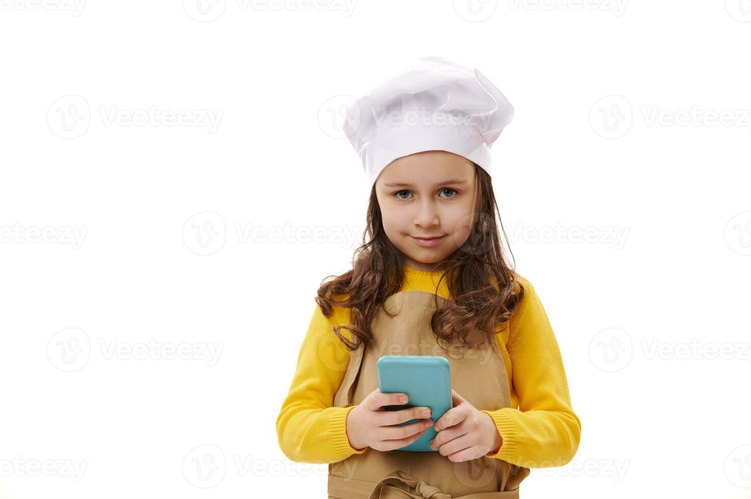 charmant bébé fille habillé comme chef confiseur, sourit à la recherche à caméra, détient une moderne téléphone intelligent plus de blanc Contexte photo