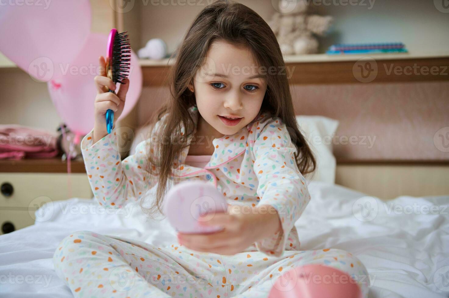 magnifique enfant fille séance sur sa lit, à la recherche à sa réflexion dans une rose petit miroir, prise se soucier de sa longue cheveux photo