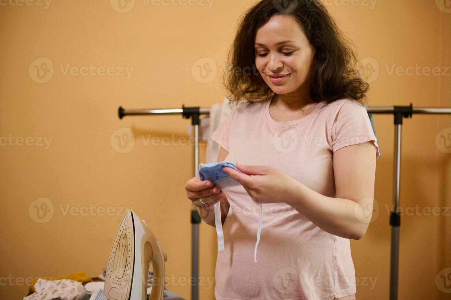 magnifique Enceinte femme souriant tandis que en portant repassé bébé casquette pour nouveau née, dans pansement pièce ou repassage pièce à Accueil photo