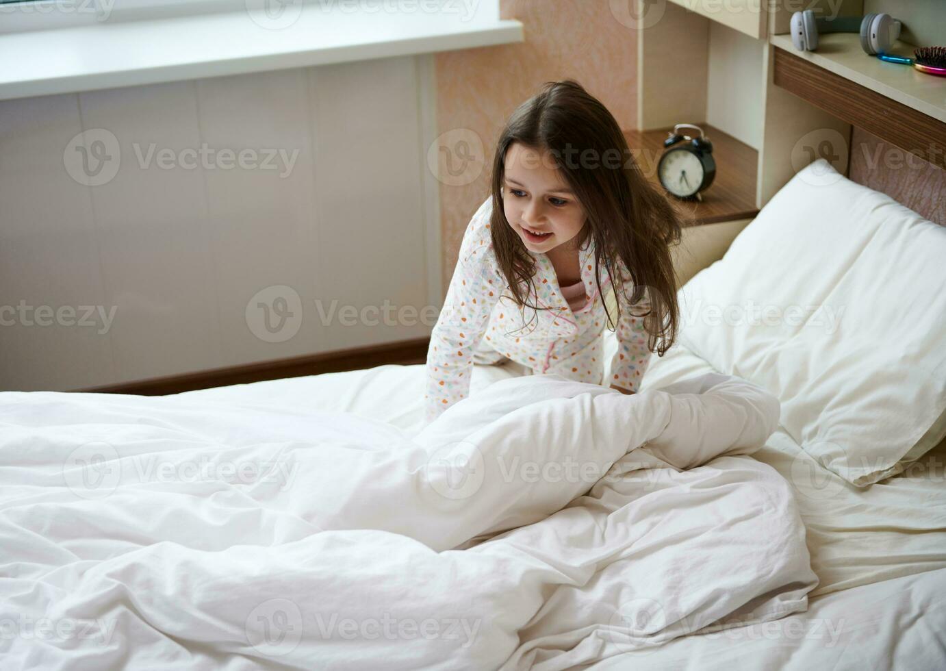charmant content caucasien enfant fille dans élégant pyjamas, veille en haut dans content ambiance et pliant sa lit dans le ensoleillé Matin photo