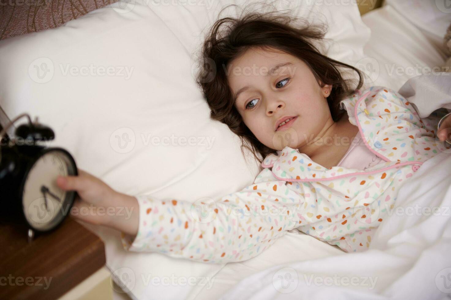 adorable enfant fille mensonge dans confortable lit, veille en haut et commutation de alarme l'horloge dans le de bonne heure Matin photo