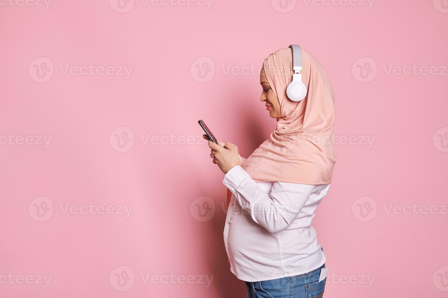 côté portrait de content arabe musulman femme dans rose hijab avec écouteurs et mobile téléphone, isolé sur Couleur Contexte photo