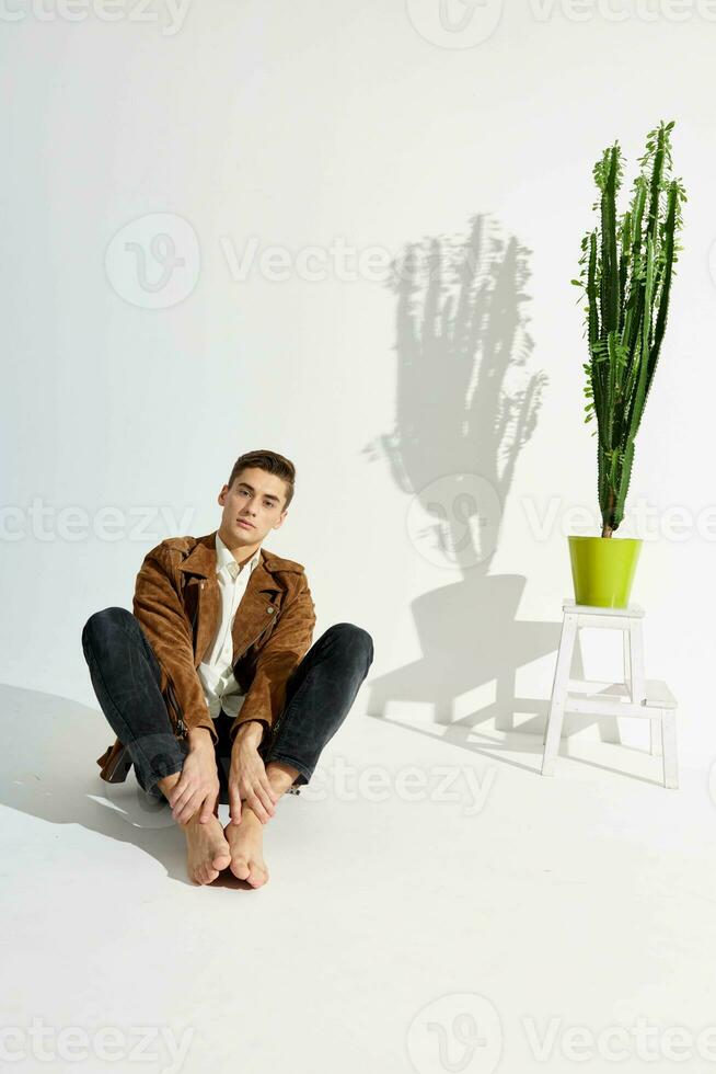 une à la mode homme dans élégant vêtements est assis sur le sol près une fleur dans une pot photo
