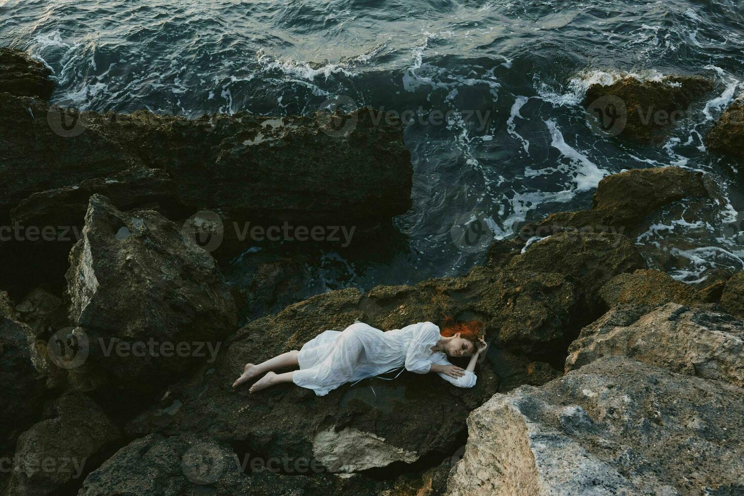 attrayant femme avec longue cheveux dans une isolé place sur une sauvage rocheux côte dans une blanc robe inchangé photo