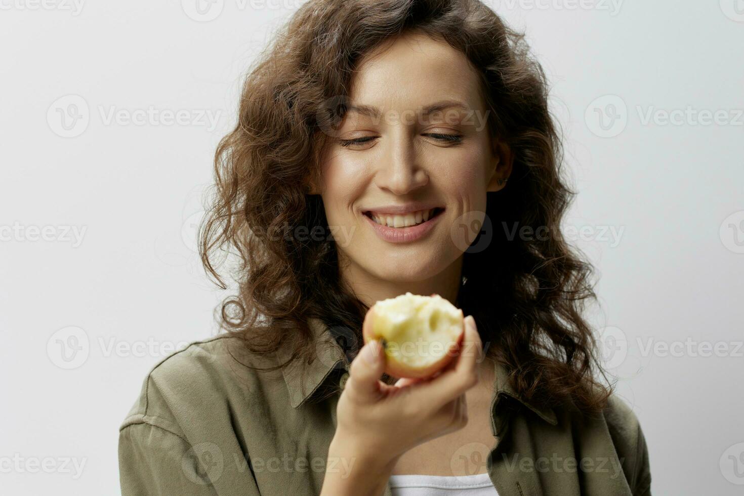 content souriant frisé magnifique femme dans décontractée kaki vert chemise manger Pomme avec un appétit considère il posant isolé sur plus de blanc Contexte. Naturel respectueux de la nature des produits concept. copie espace photo