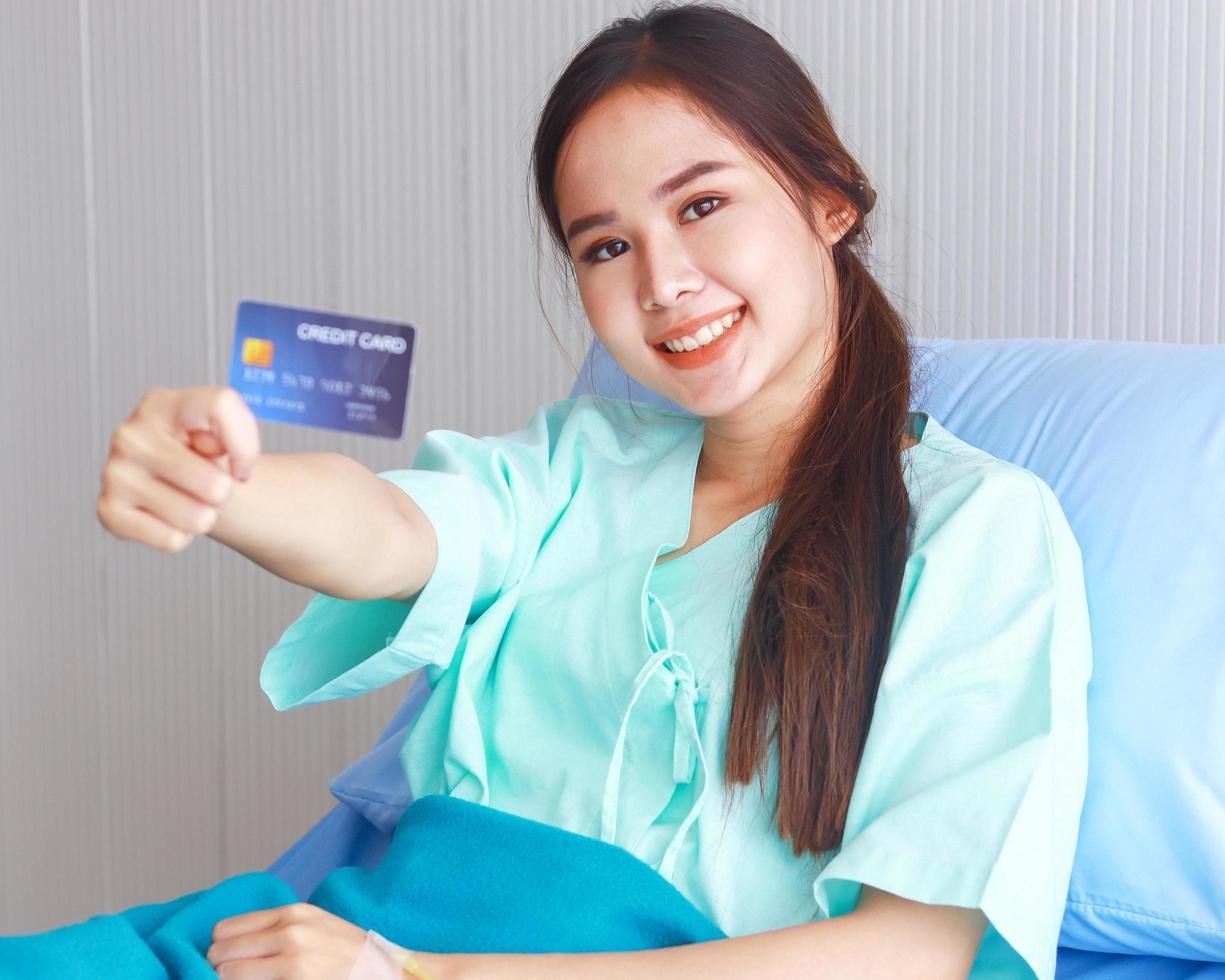 Belle femme asiatique avec une carte de crédit est assise dans un lit d'hôpital avec un visage souriant photo