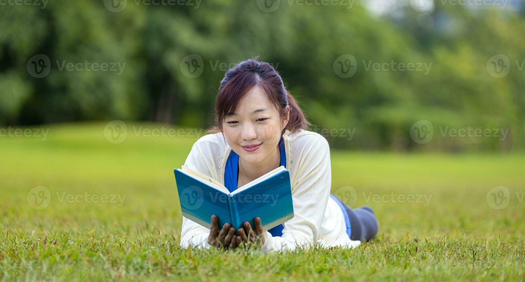 asiatique femme est mensonge vers le bas dans le herbe pelouse à l'intérieur le Publique parc en portant livre dans sa main pendant été pour en train de lire et éducation concept photo
