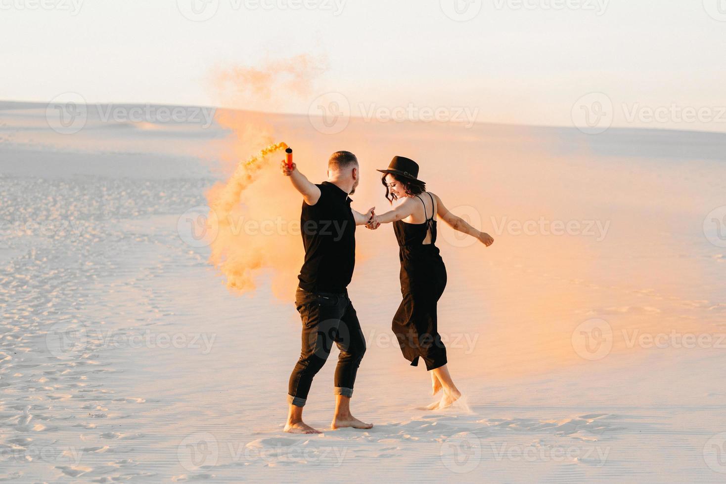 mec et une fille en vêtements noirs étreignent et courent sur le sable blanc avec de la fumée orange photo