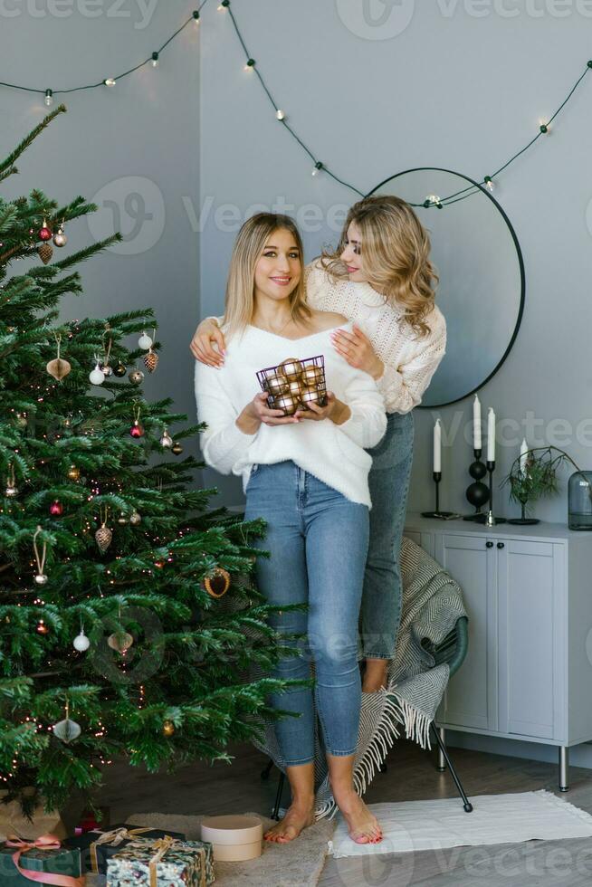 deux magnifique blond sœurs. ayant amusant, avoir prêt pour le fête, décorer le Noël arbre. une brillant fête de meilleur copains habillé dans chaud hiver chandails photo