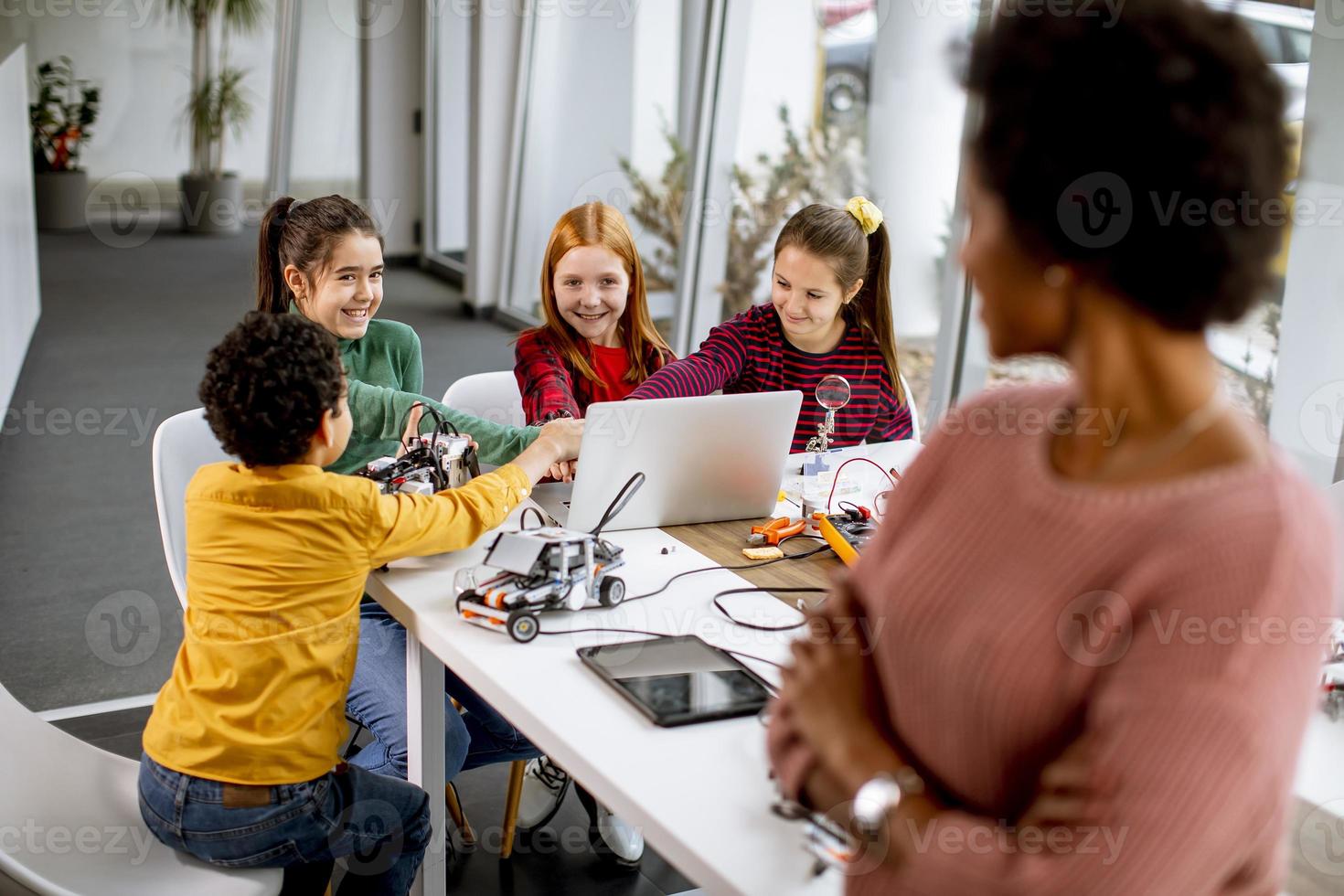 Des enfants heureux avec leur professeur de sciences afro-américaine avec des jouets électriques et des robots de programmation pour ordinateur portable à la classe de robotique photo