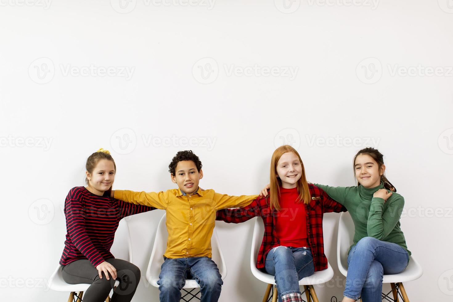 Portrait de mignons petits enfants en jeans assis sur des chaises contre le mur blanc photo