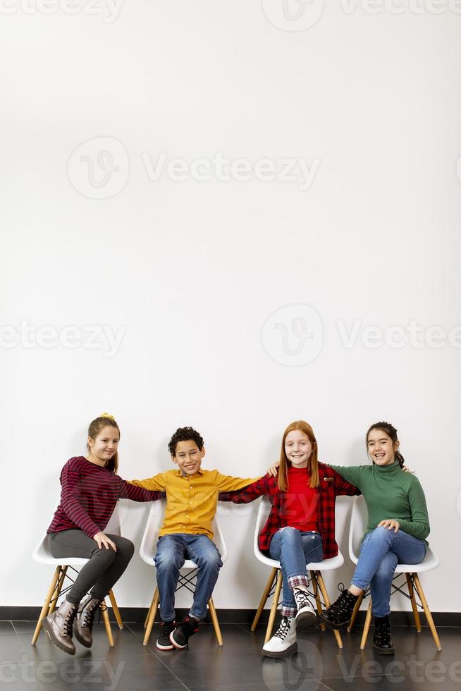 Portrait de mignons petits enfants en jeans assis sur des chaises contre le mur blanc photo