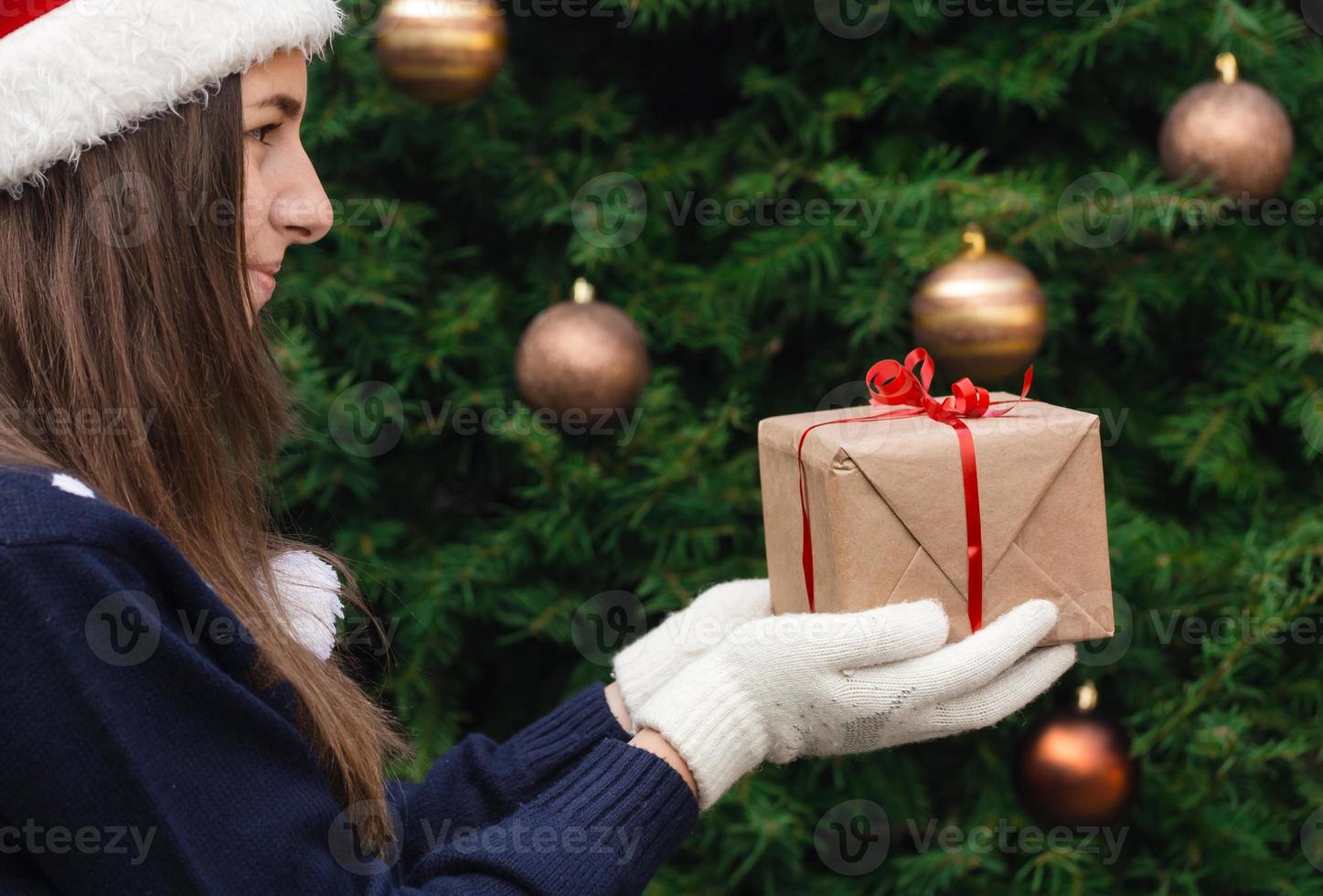 Une jeune fille portant un chapeau de père Noël donne un cadeau en papier kraft avec un ruban rouge photo