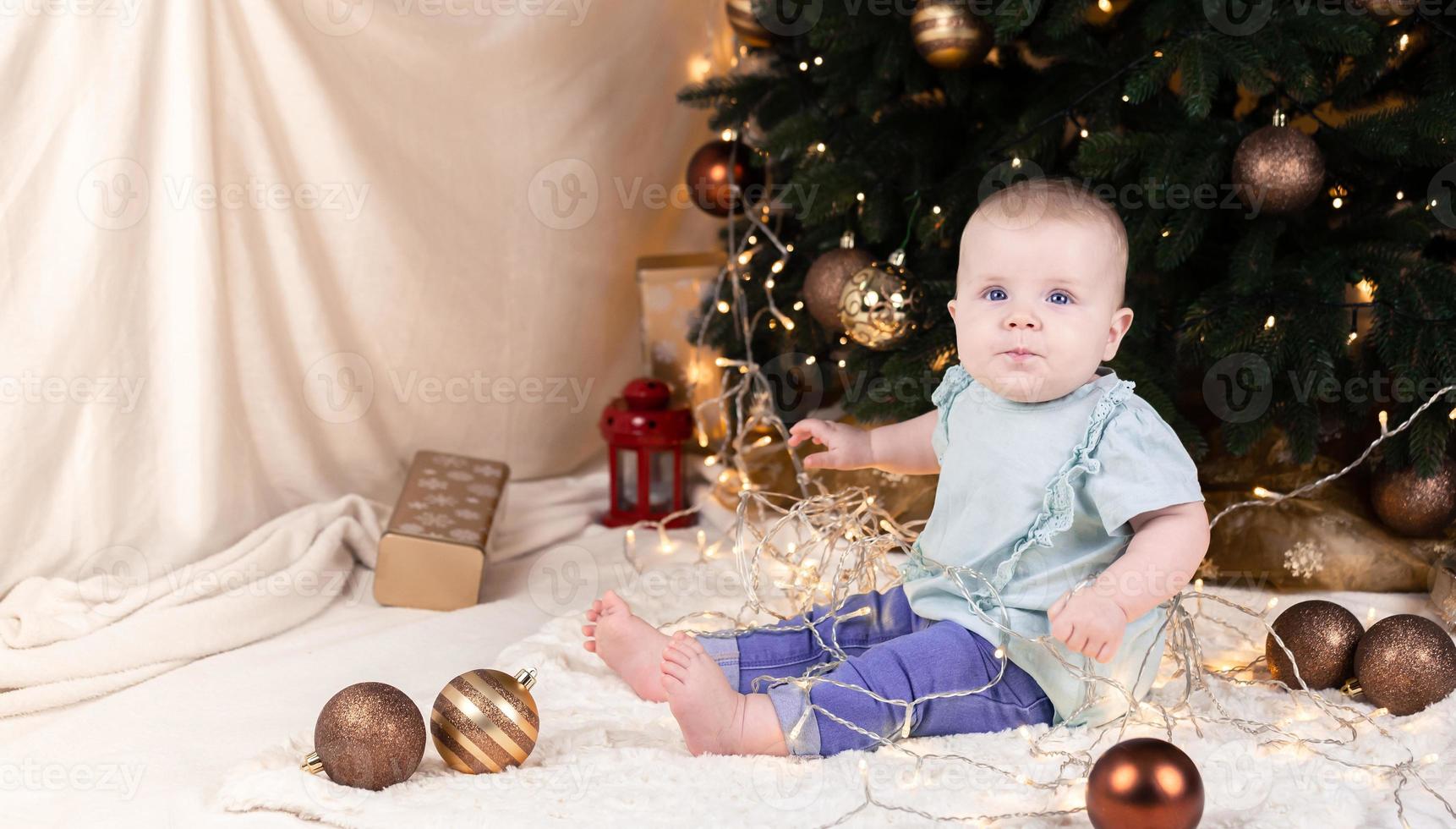 un bébé en jean est assis près d'un arbre de Noël et joue avec une guirlande dans laquelle il est empêtré photo
