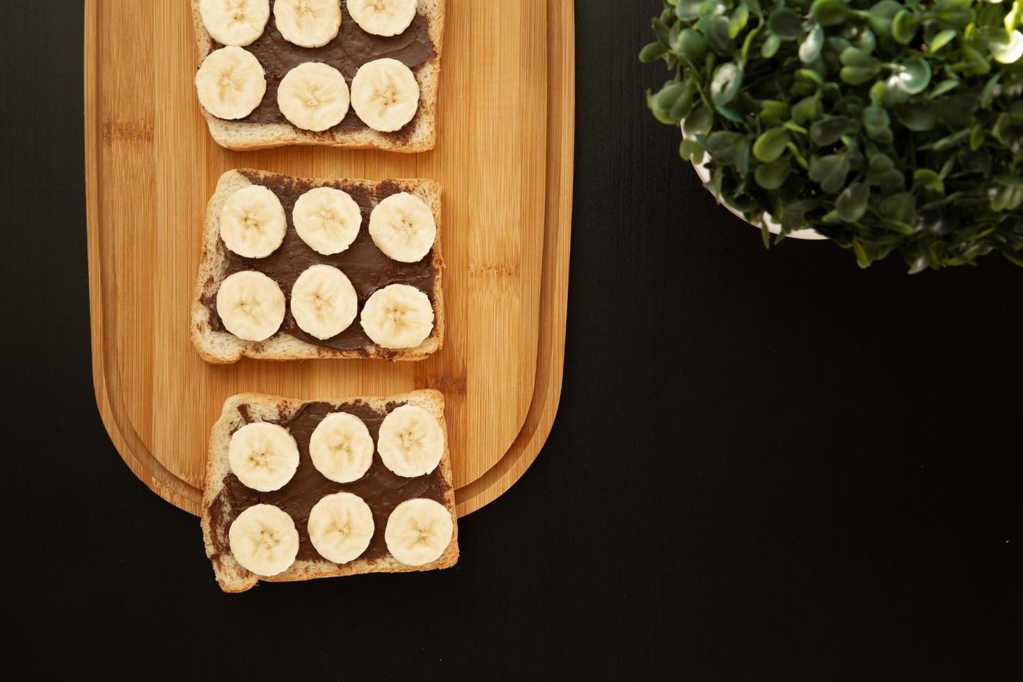 Trois toasts de pain blanc à la banane enduits de beurre au chocolat sur une planche à découper sur un fond sombre photo