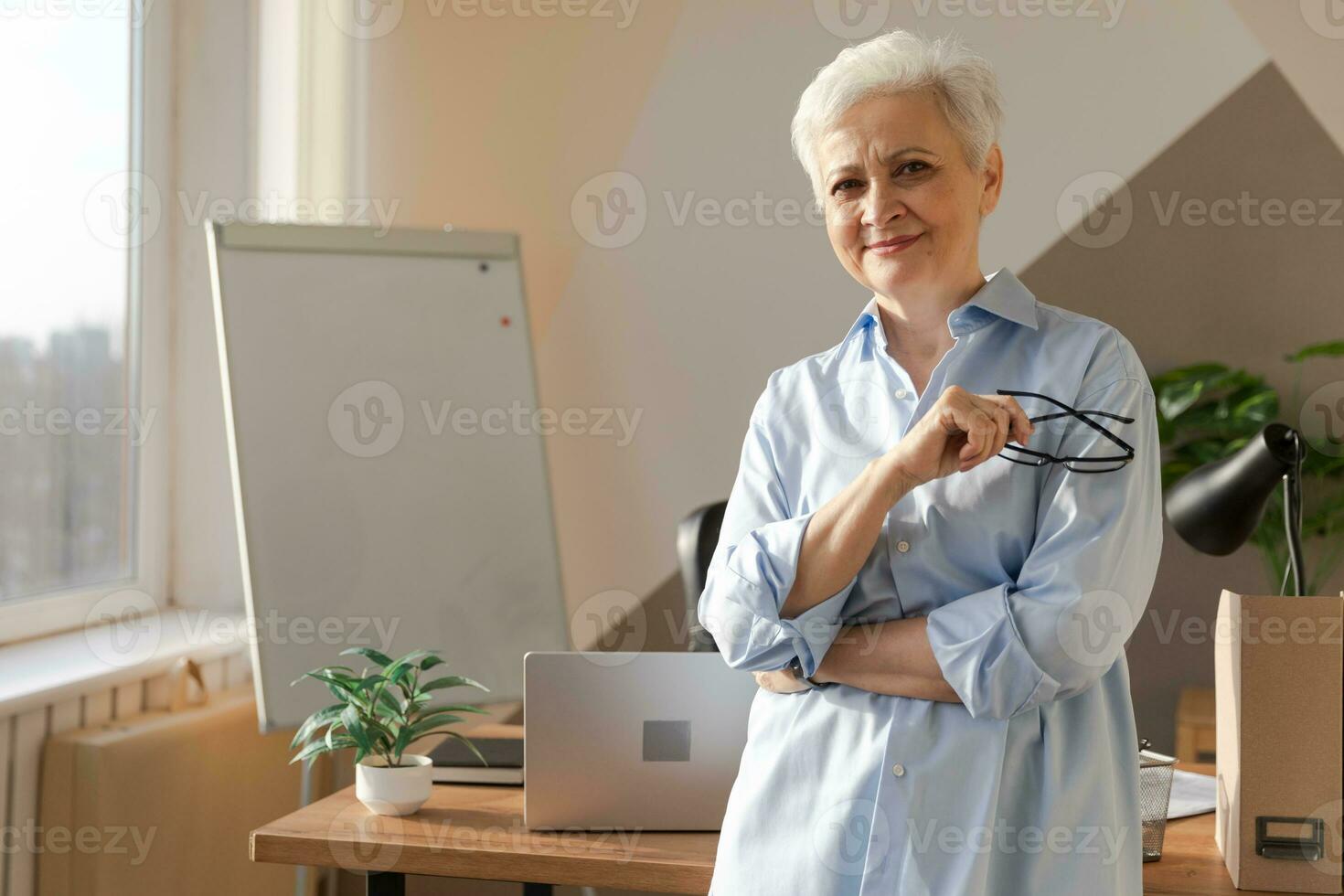 portrait de sur de soi élégant européen milieu vieilli Sénior femme à lieu de travail. élégant plus âgée mature Années 60 gris aux cheveux Dame femme d'affaires dans moderne bureau. patron chef prof professionnel ouvrier. photo