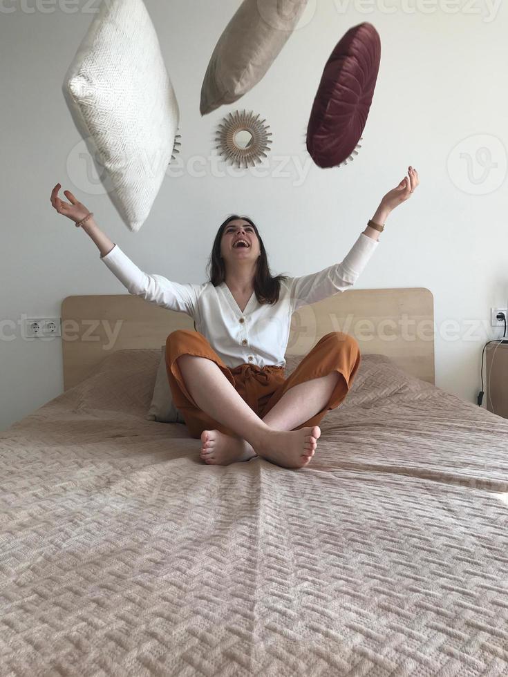 fille joyeuse assise sur le lit en lançant l'oreiller et souriant photo