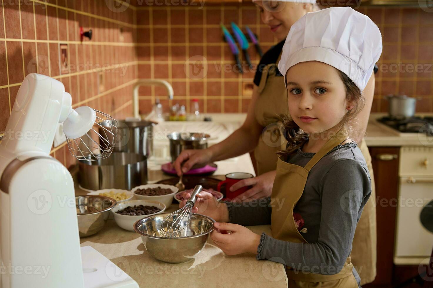 charmant enfant fille dans du chef chapeau et tablier, aide sa maman à faire une de fête gâteau mélange fouetté crème avec fondu Chocolat photo