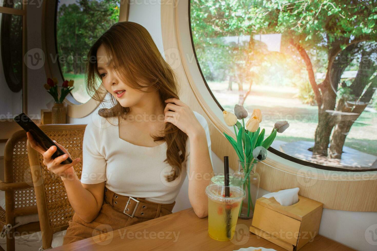 Jeune adulte content asiatique femme en utilisant intelligent téléphone pour social médias application, achats en ligne, transfert argent, financier, message dans café. Contexte avec fenêtre et chaud lumière du soleil sur hiver saison. photo