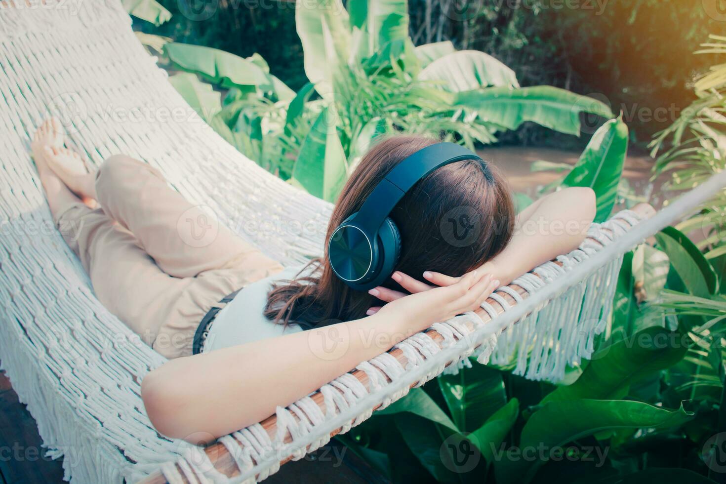 Jeune détendu femme écoute à la musique avec écouteurs sur blanc hamac dans jardin à maison. Matin Soleil. lent vie, fin de semaine loisir activité. quarantaine et soi isolement période. photo