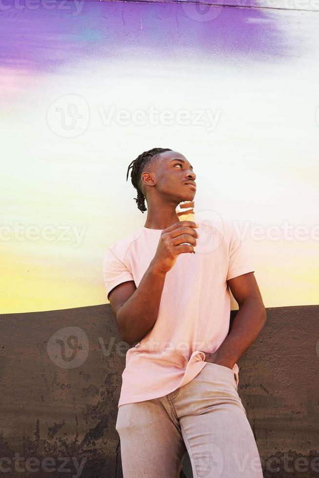 Jeune bel homme noir porte une chemise rose et mange un cornet de crème glacée en été sur un mur peint comme un lever de soleil ou une journée ensoleillée photo