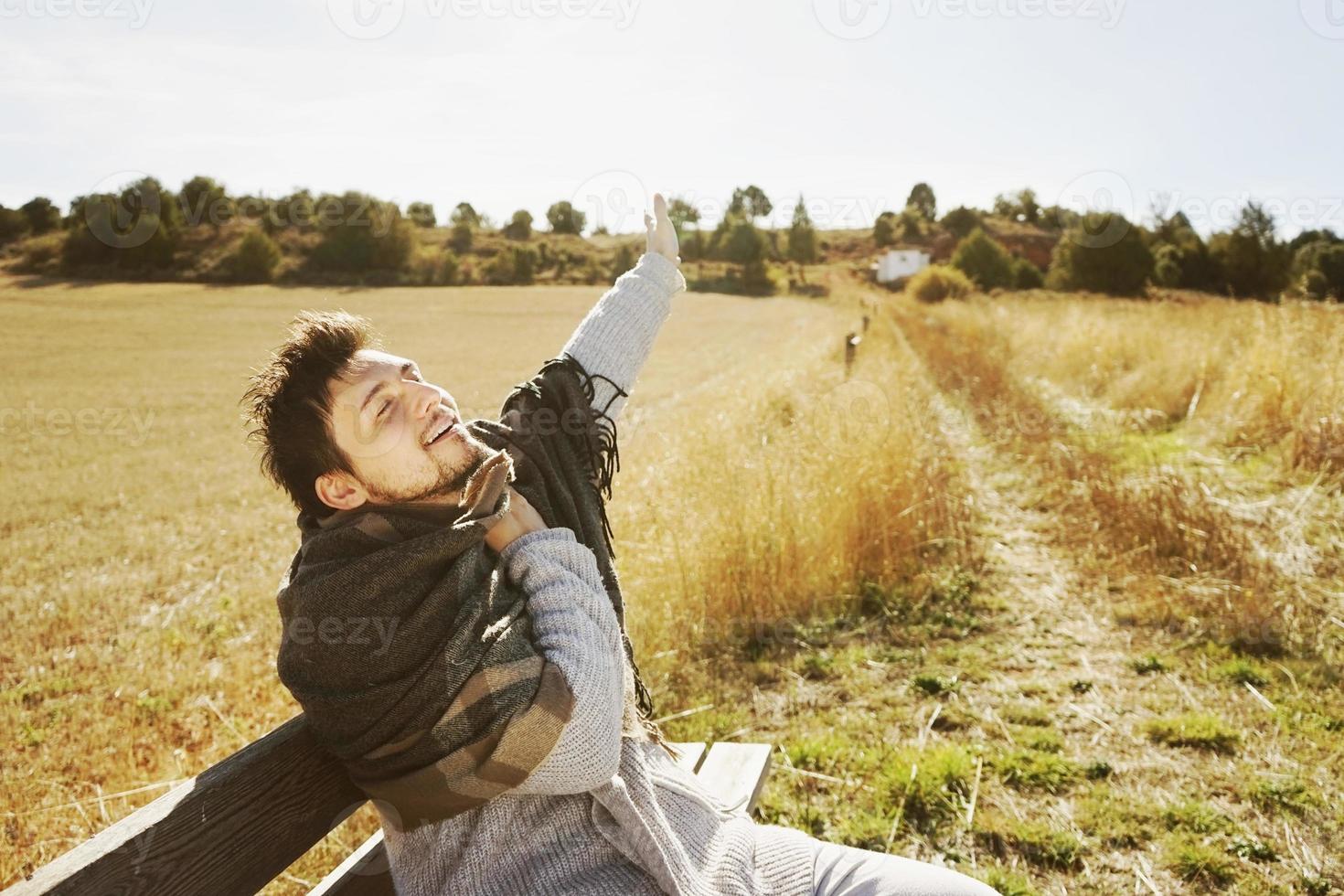 Un jeune homme aux bras étendus et une écharpe chaude profitant du soleil d'automne du matin dans un champ jaune avec le contre-jour du ciel bleu photo