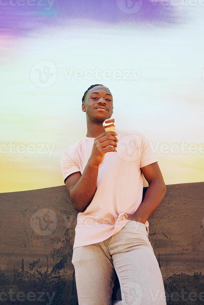 Jeune bel homme noir porte une chemise rose et mange un cornet de crème glacée en été sur un mur peint comme un lever de soleil ou une journée ensoleillée photo