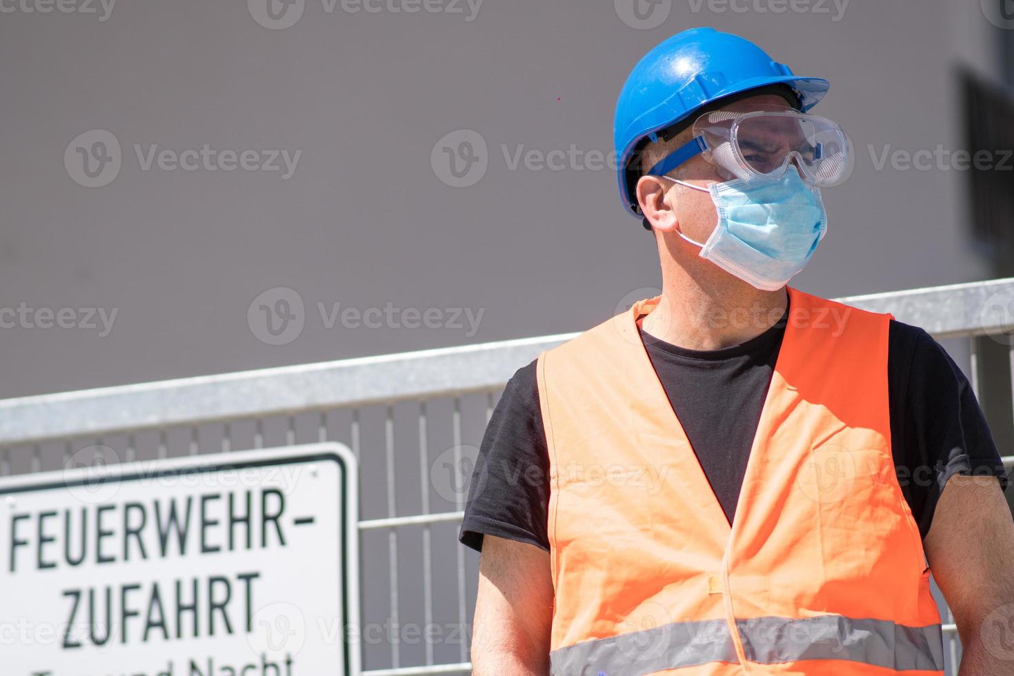 Ouvrier du bâtiment portant un casque bleu, un gilet réfléchissant et un masque chirurgical de protection photo