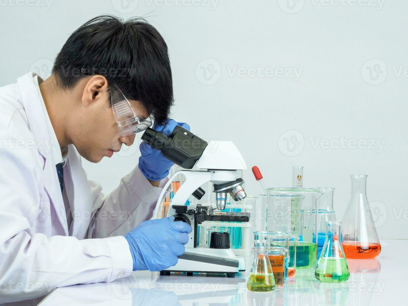 asiatique Masculin étudiant scientifique portant une médecins robe dans le laboratoire, à la recherche à le résultats par une microscope. sur une table dans une scientifique recherche laboratoire avec tester tubes sur une blanc table photo