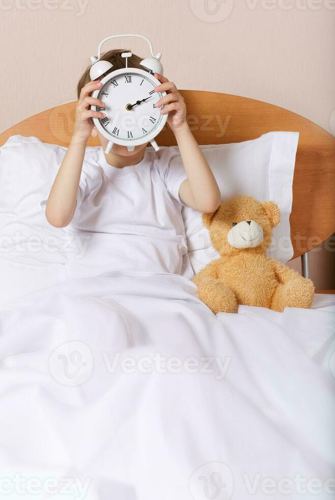 garçon est séance dans le lit avec gros alarme l'horloge photo