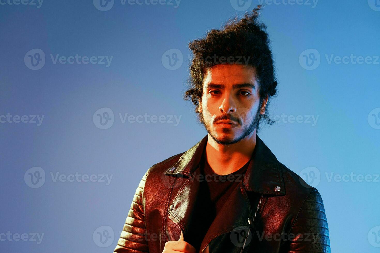 portrait de une élégant homme avec frisé cheveux sur une bleu Contexte multinational, coloré lumière, noir cuir veste s'orienter, moderne concept. photo