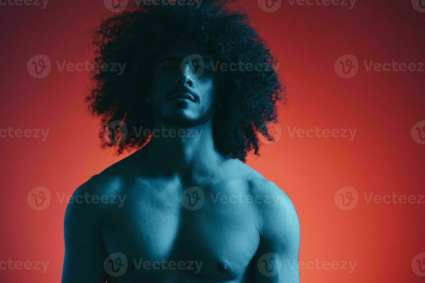 portrait de mode homme avec frisé cheveux sur rouge Contexte avec élégant lunettes, multinational, coloré lumière, noir cuir veste s'orienter, moderne concept, sexy corps. photo