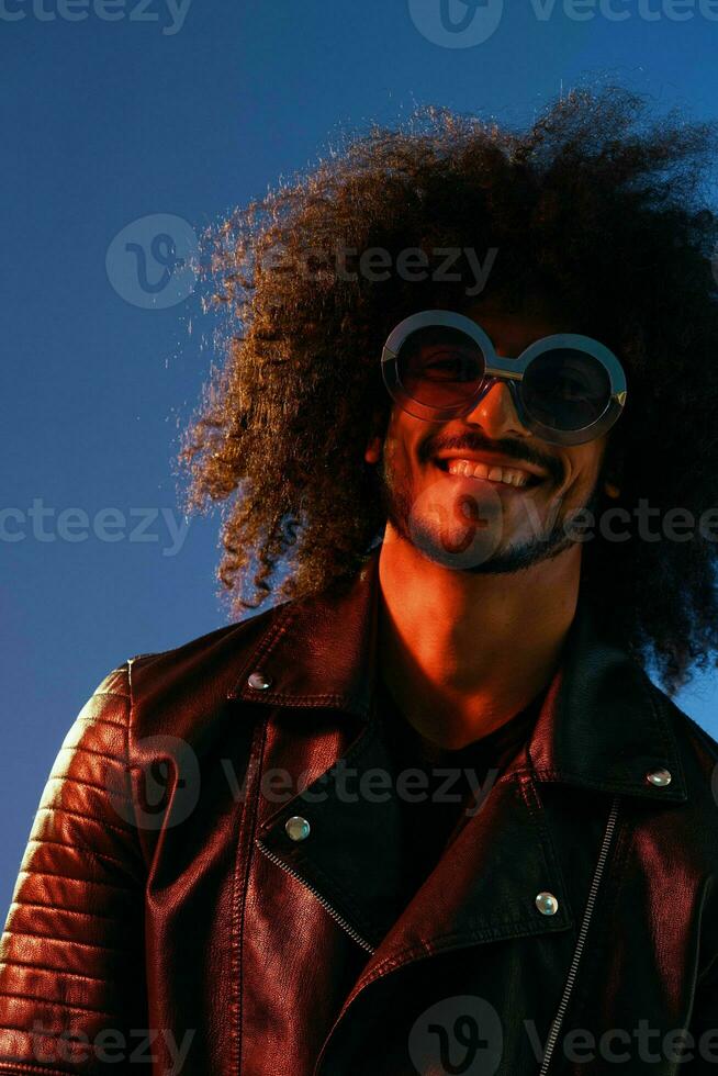 portrait de une élégant homme avec frisé cheveux avec des lunettes sourire sur bleu Contexte multinational, Couleur lumière, noir cuir veste s'orienter, moderne concept. photo