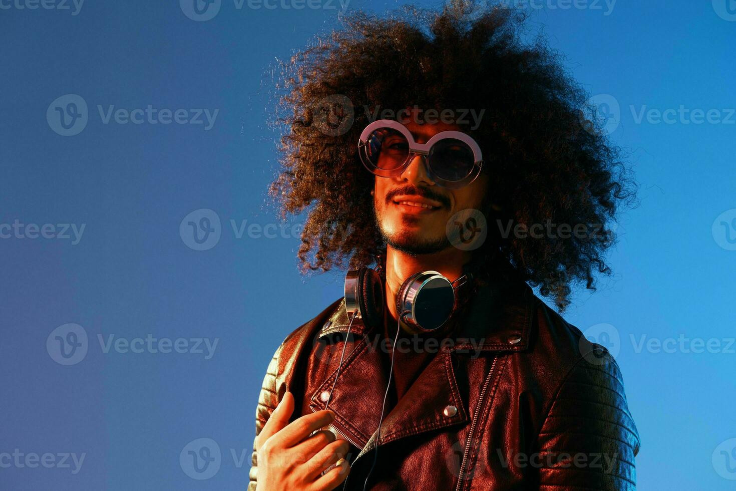 portrait de une élégant homme avec frisé cheveux avec des lunettes et écouteurs sur une bleu Contexte multinational, coloré lumière, noir cuir veste s'orienter, moderne concept. photo