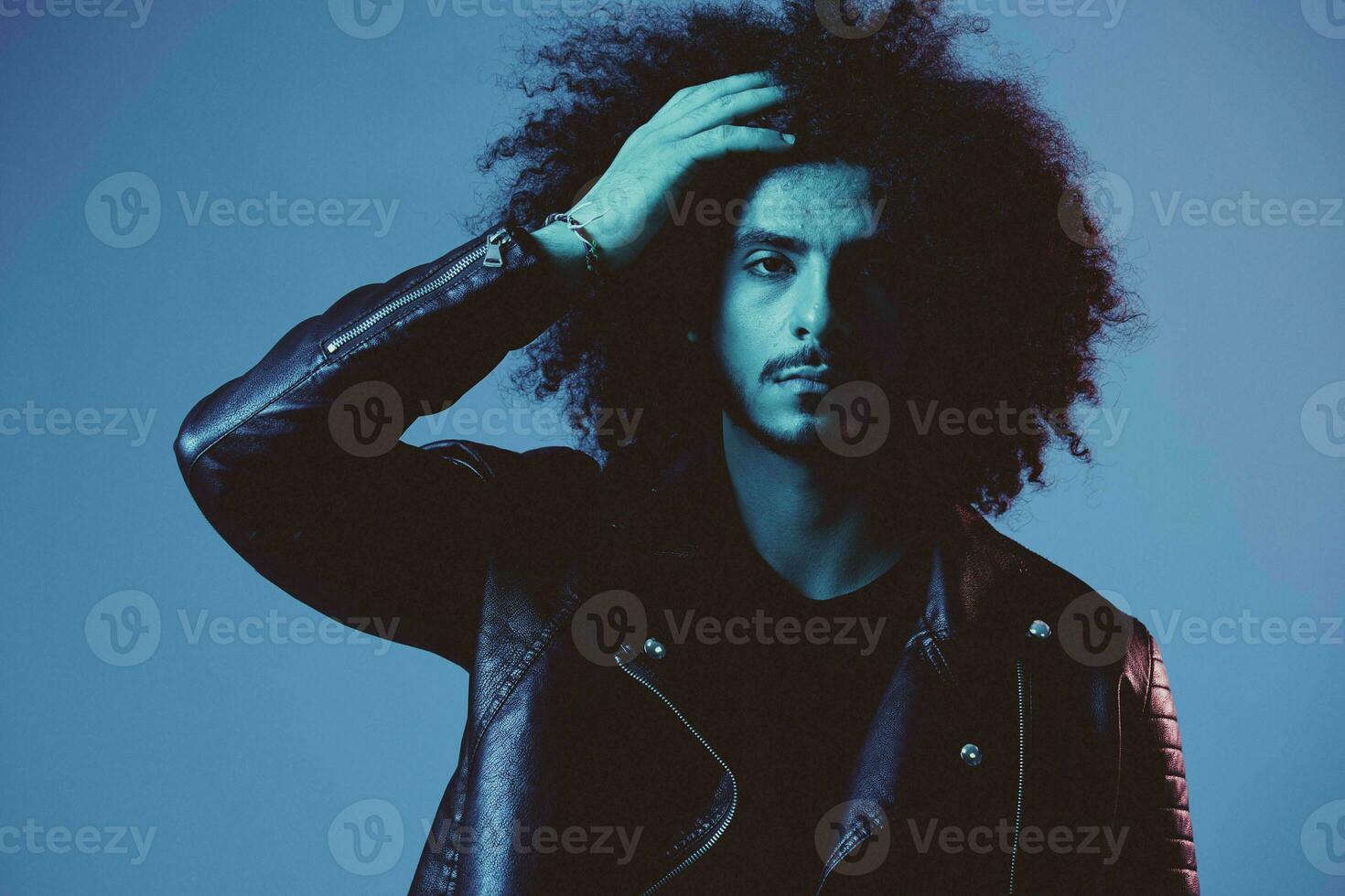 portrait de mode homme avec frisé cheveux sur bleu Contexte multinational, coloré lumière, noir cuir veste s'orienter, moderne concept. photo