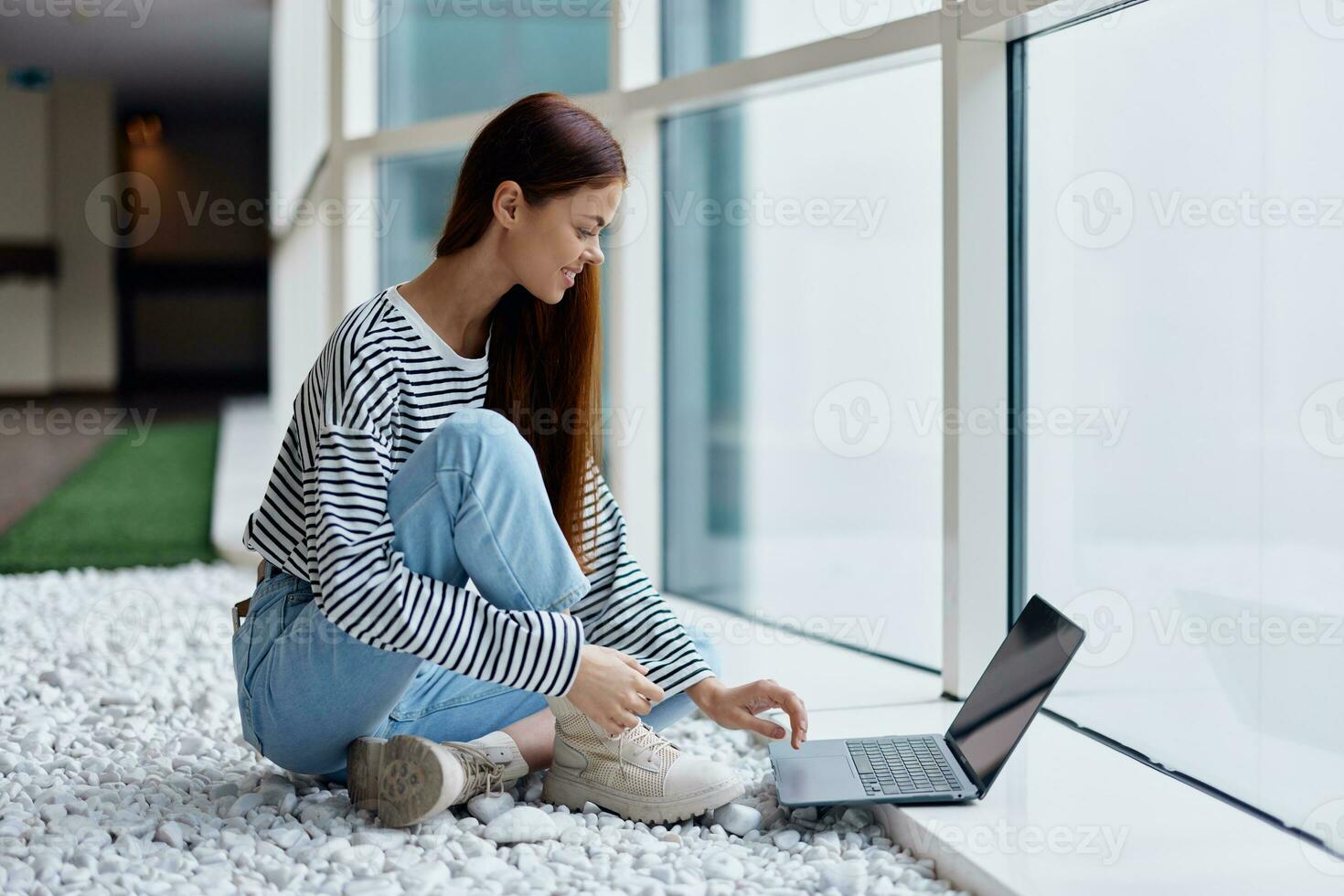 une femme souriant avec les dents est assis avec une portable par le grand les fenêtres de une bâtiment dans le ville parlant via vidéo lien par une ordinateur, le concept de travail et loisir dans le gros ville photo