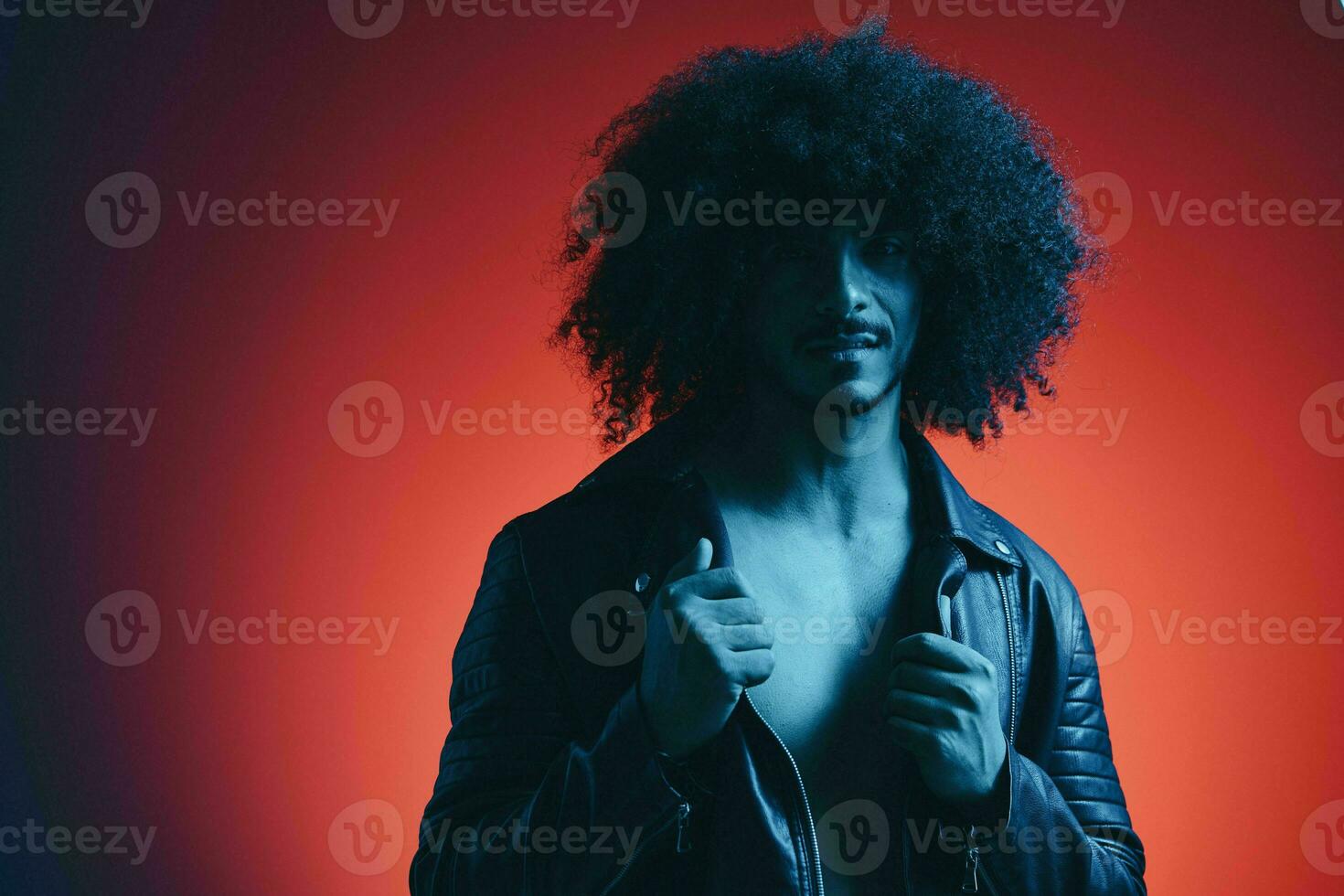 portrait de mode homme avec frisé cheveux sur rouge Contexte multinational, coloré lumière, noir cuir veste s'orienter, moderne concept. photo