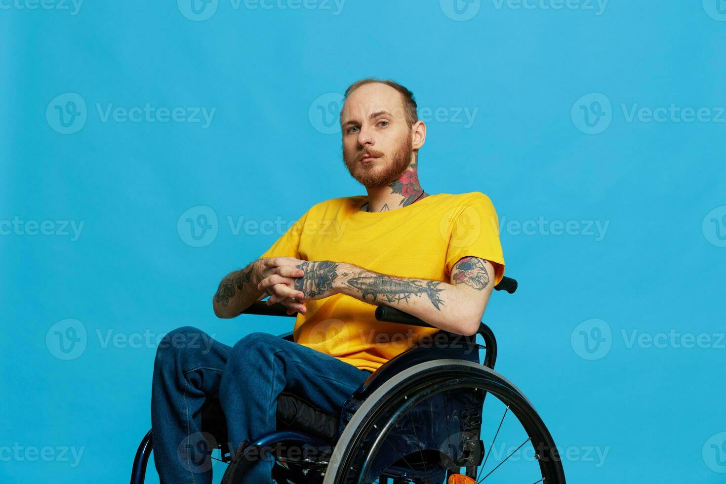 une homme dans une fauteuil roulant problèmes avec le musculo-squelettique système regards à le caméra dans une T-shirt avec tatouages sur le sien bras est assis sur une bleu studio arrière-plan, plein vie, réel personne, santé concept photo