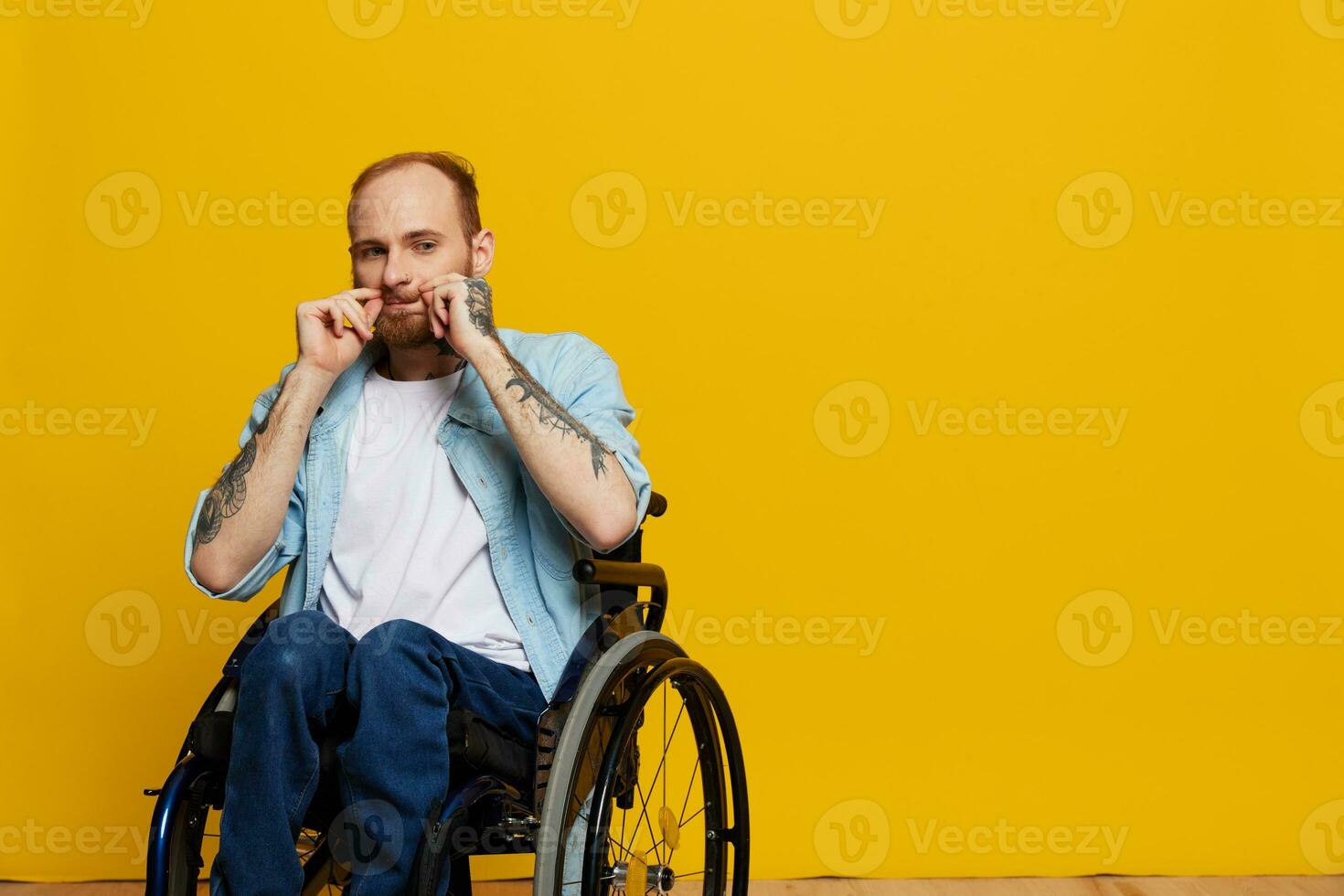une homme dans une fauteuil roulant regards à le caméra, avec tatouages sur le sien bras est assis sur une Jaune studio arrière-plan, le concept de santé est une la personne avec handicapées photo