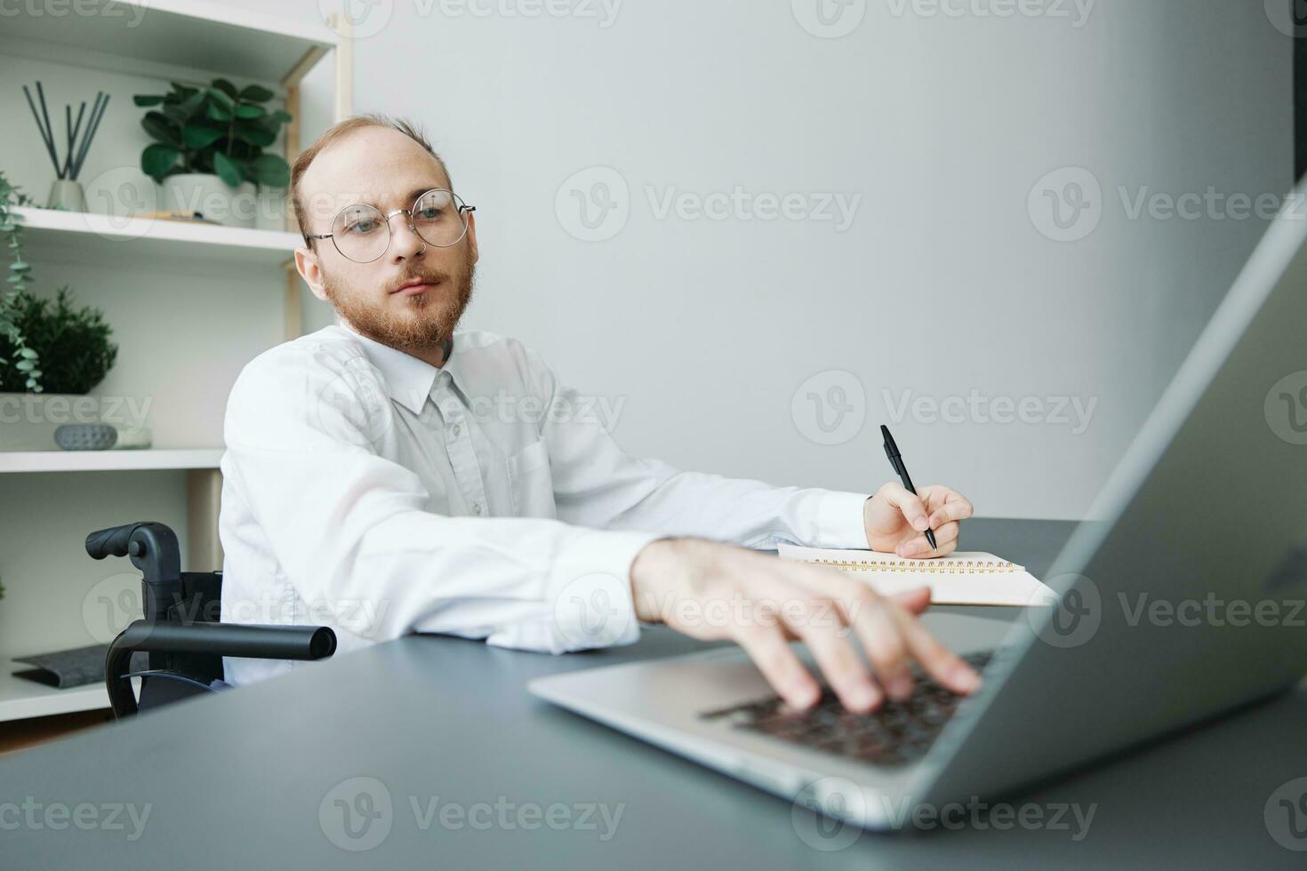 une homme fauteuil roulant homme d'affaire dans le Bureau travaux à une ordinateur portable, l'écriture vers le bas des idées et une plan dans une carnet de notes, prévenance, l'intégration dans société, le concept de travail une la personne avec handicapées photo